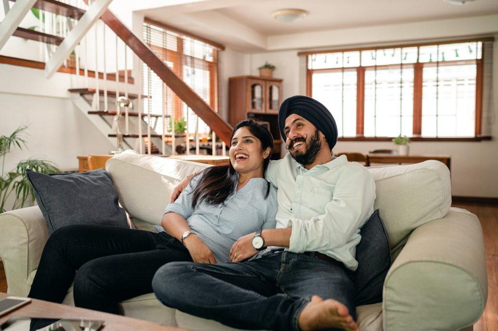 Happy young Indian man in turban and positive woman in casual clothes embracing and laughing while watching comedy movie sitting on sofa